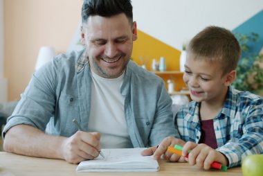 Cheerful dad is drawing funny pictures for happy child having fun sitting at desk at home enjoying leisure time indoors. Youth, creative people and arts concept.